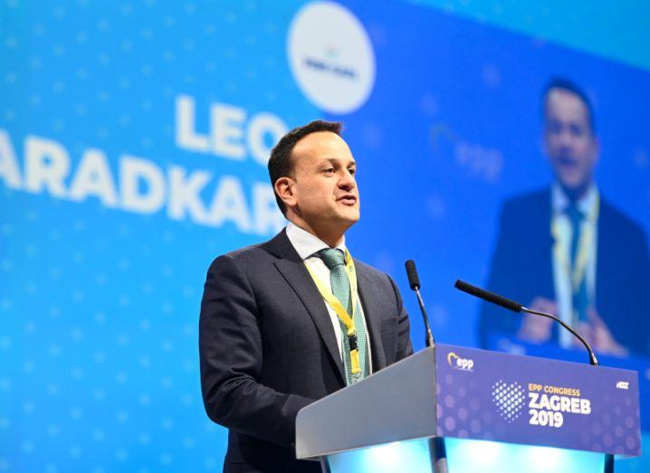 A man standing on a stage in front of a podium, with two microphones in front of him and a blue background. He is Leo Varadkar.