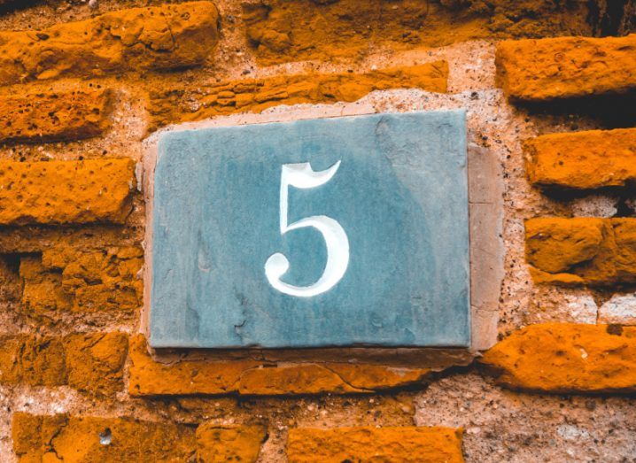 A grey slate tile with the number five embossed in white on an uneven orange brick wall.