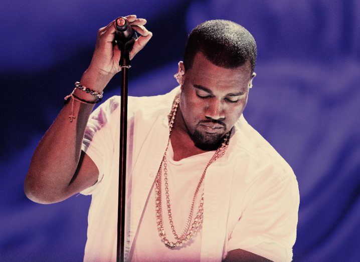The rapper Kanye West wears a white outfit and gold chains. He is holding a microphone stand and looking down.