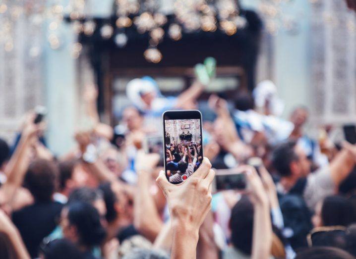 A person holding a smartphone, recording a video of an event in the background.