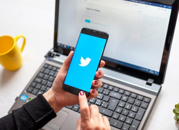 A person holding a smartphone with the Twitter logo on the screen. A laptop is behind the phone that has a Twitter log-in page on the screen. There is a yellow cup to the left of the laptop, resting on a white table.