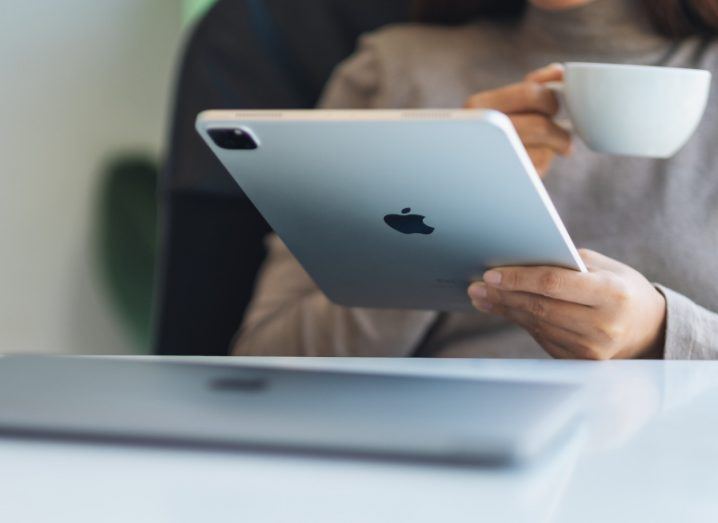A person holding an iPad with a cup in their other hand. An Apple mac is laying on a table in front of the person.