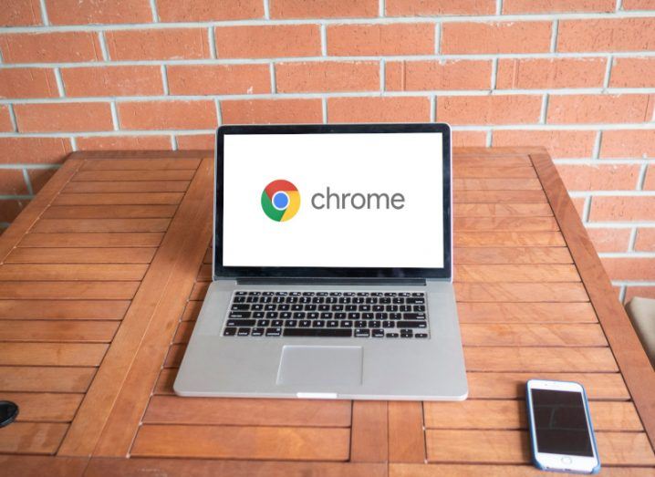 A Google Chrome logo on a laptop screen. The laptop is resting on a wooden table next to a smartphone with a brick wall behind it.