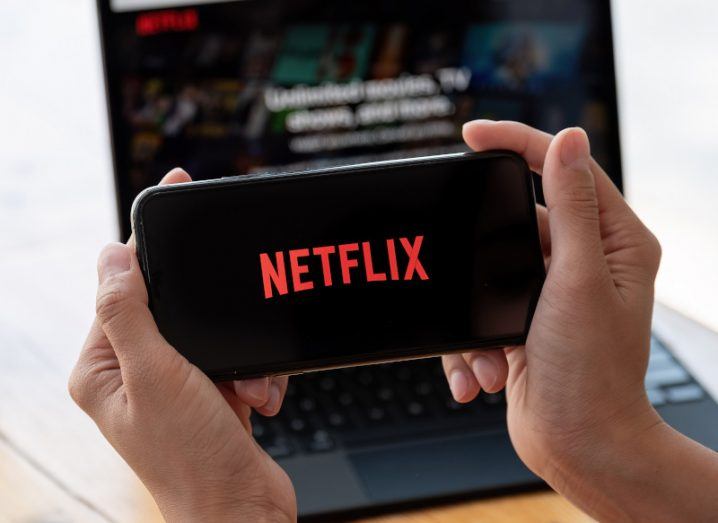 A smartphone with the Netflix logo on the screen being held in a person's hands, with a laptop in the background. The Netflix logo is also on the laptop screen.