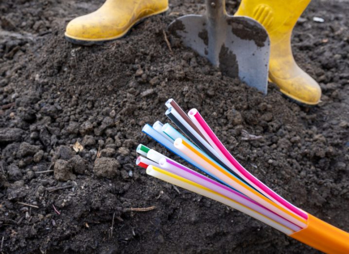 A pair of yellow work boots frame a shovel being dug into the ground in order to lay a fibre broadband cable, which is pictured in the foreground.