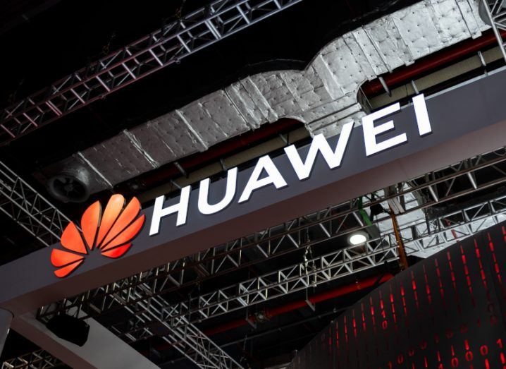 Huawei logo and name on a metal bar near the ceiling of a large building, with red lights visible behind it.