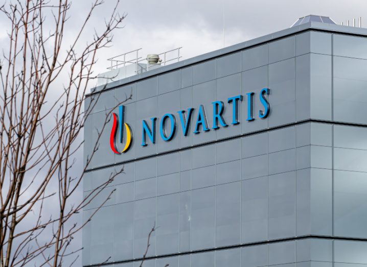 A building with the Novartis logo on the side, with a grey cloudy sky above it and a tree with no leaves to the side of the image.