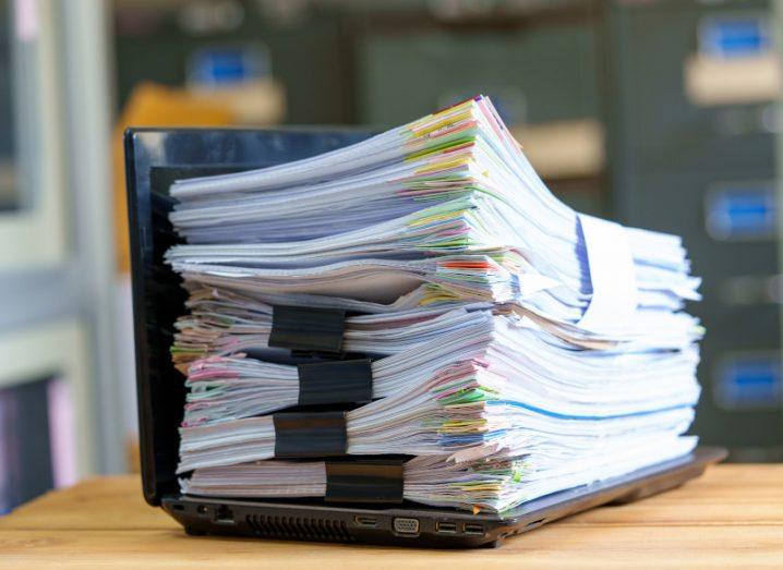 A batch of papers on top of a laptop, resting on a desk with an office background.