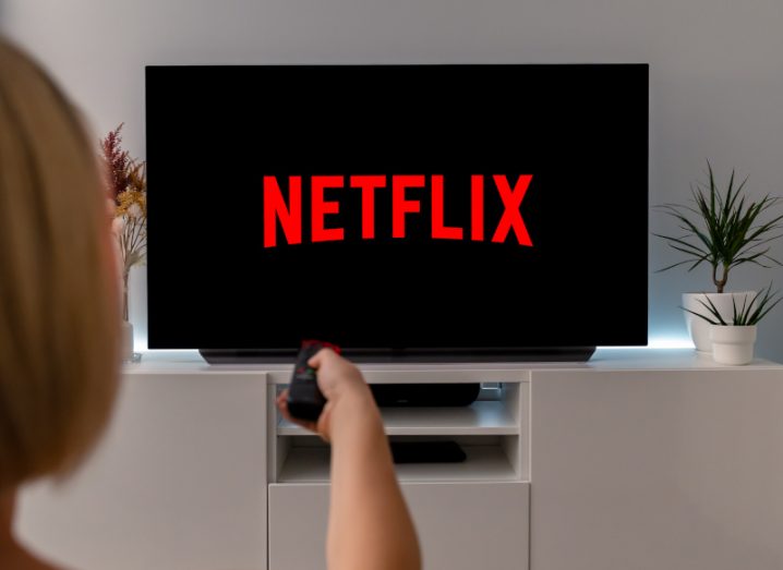 Netflix logo on a TV screen, with a person pressing a remote in front of the TV. The device is resting on a white table next to a small houseplant.