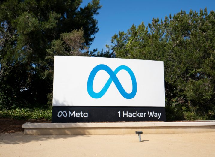 Meta company logo on a white billboard with the address of 1 Hacker Way written on the bottom right. The billboard is in front of trees, with a clear blue sky overhead.