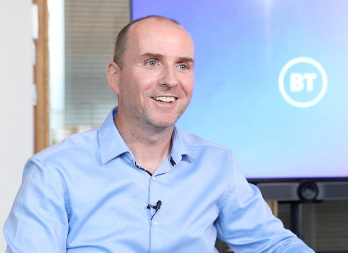 A man in a light blue shirt smiles, looking slightly off camera. A BT logo is in the background.