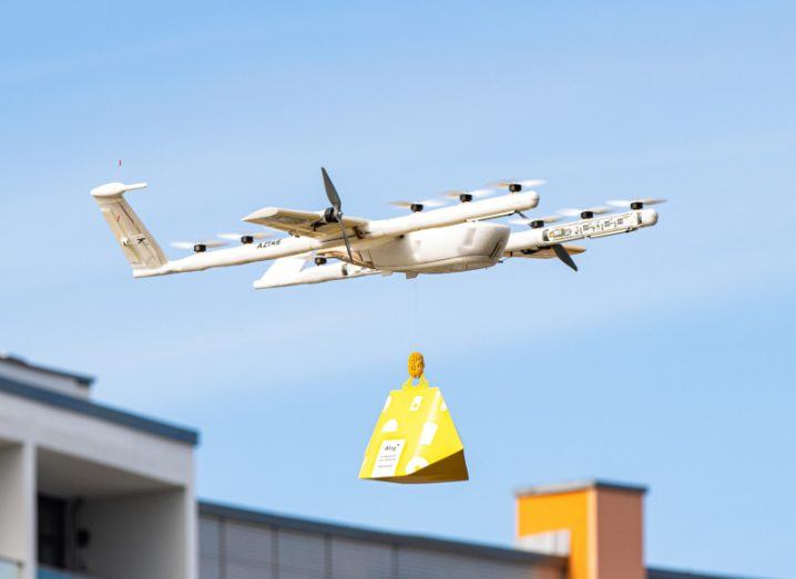 Image of a Wing drone with a building and blue sky in the background. The drone has a delivery package connected under it.