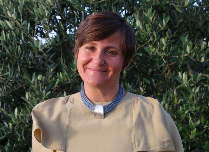 A woman with short dark hair smiles at the camera against a backdrop of shrubbery. She is Gaia Scalabrino.