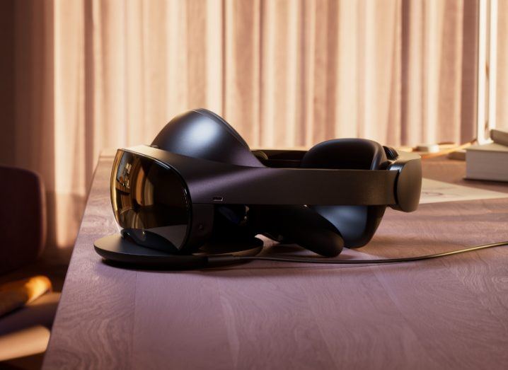 Image of a virtual reality headset resting on a wooden table with curtains in the background. It is the Meta Quest Pro.