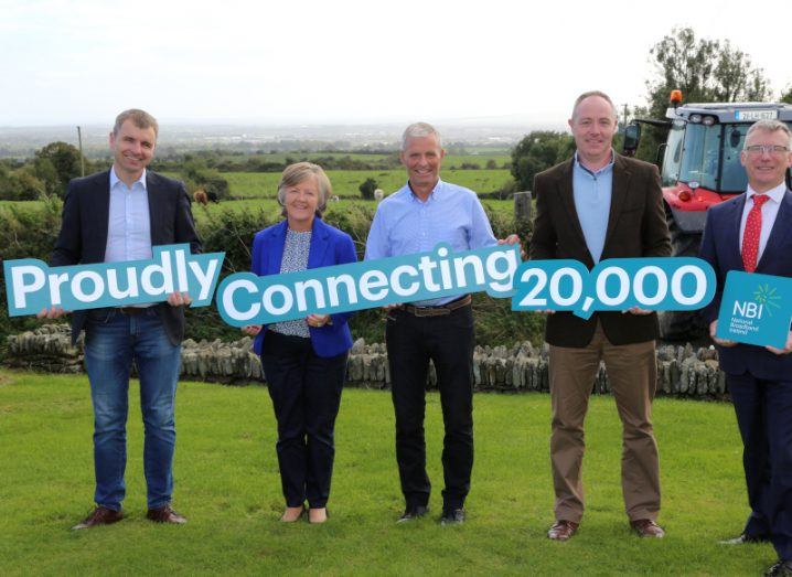 Four men and a woman standing in a field, holding signs that say 'Proudly Connecting 20,000'. The signs are related to the National Broadband Plan roll-out, being managed by National Broadband Ireland (NBI).