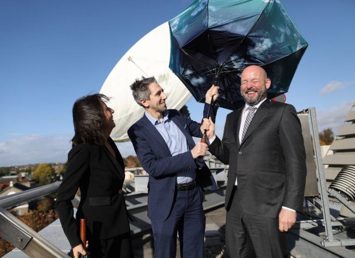 Two men and a woman standing in front of a satellite dish, with the two men holding an umbrella that has gone inside out. A blue sky is visible in the background.