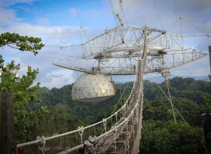 Image of a bridge leading to the Arecibo Observatory, with trees and a blue sky with some grey clouds visible in the background.