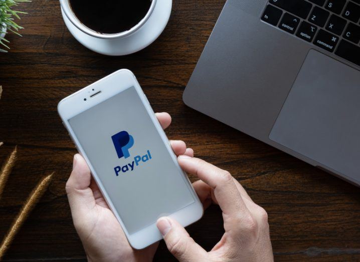 A person holding a white iPhone with the PayPal logo on the screen. There is a coffee and a laptop on the table nearby.