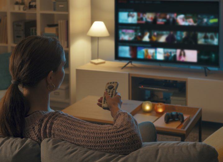 A woman sits on a couch and holds a remote control pointed at a TV with lots of TV show thumbnails visible.