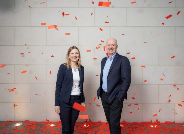 A man and a woman smiling next to each other with red confetti falling around them and a white tiled wall behind them. They are the CEOs of Virgin Media Ireland and Vodafone Ireland.