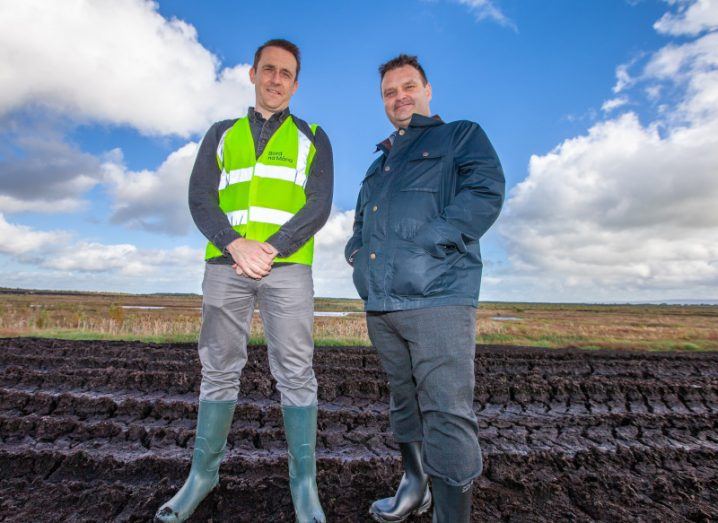 Two men standing together in the middle of a field, with a blue sky with grey clouds in the background. One of the men is wearing a high visibility vest. They are part of Bord na Móna and Esri Ireland.