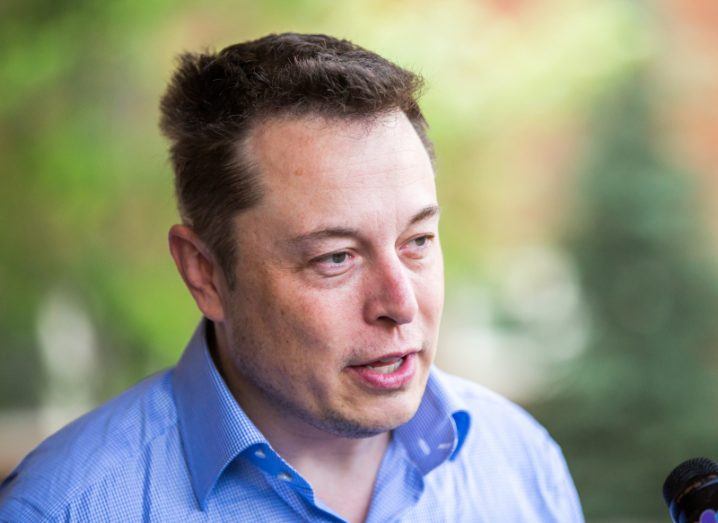 A man in a blue shirt speaking in front of a microphone. He is Elon Musk.