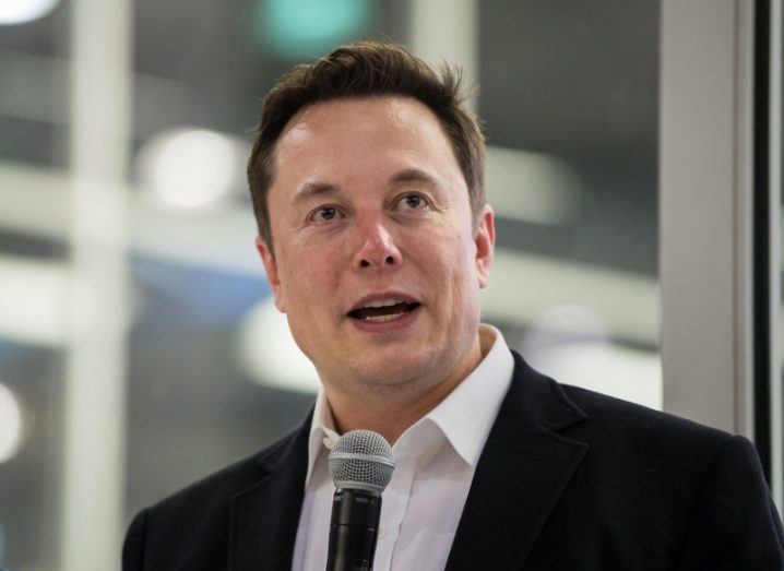 A man in a black suit speaking in front of a microphone. He is Elon Musk, owner of companies such as Twitter and SpaceX.