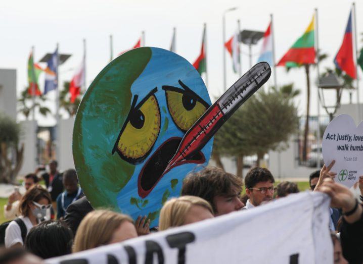 A crowd holding up signs with country flags in the background. The center of the image has an illustration of the Earth with a face that looks unwell, with a thermometer in its mouth. Taken during the COP27 conference.