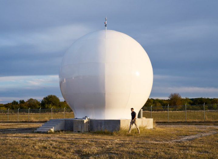A ground station in the middle of a field, with a person walking next to it. The station is owned by Globalstar and is used for Apple's Emergency SOS feature for iPhone 14 models.