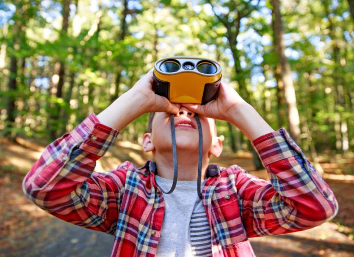 A boy looking up at the sky with a pair of binoculars as he walks through a forest.