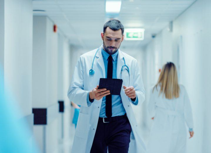 A doctor walking down a hospital hallway, holding a tablet in his hand. There is another doctor behind him walking the opposite direction.