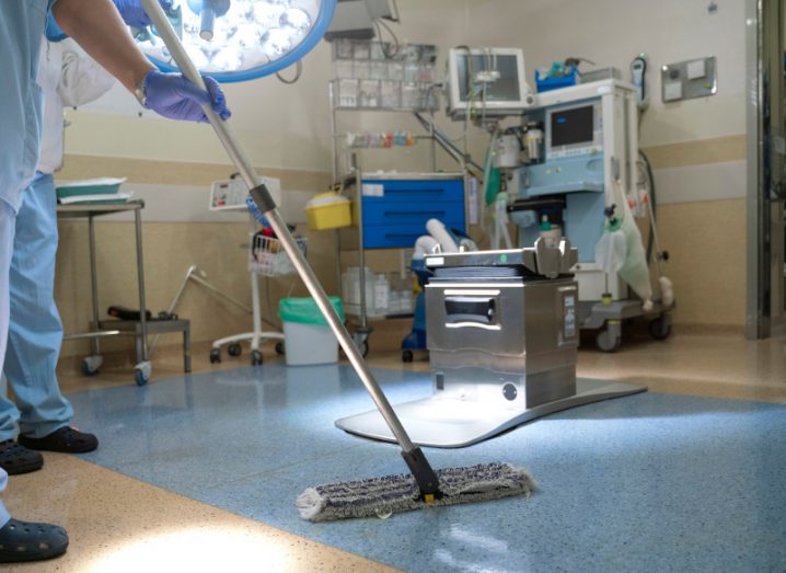 A hospital staff worker cleaning an operating room with a mop, with medical equipment in the background.