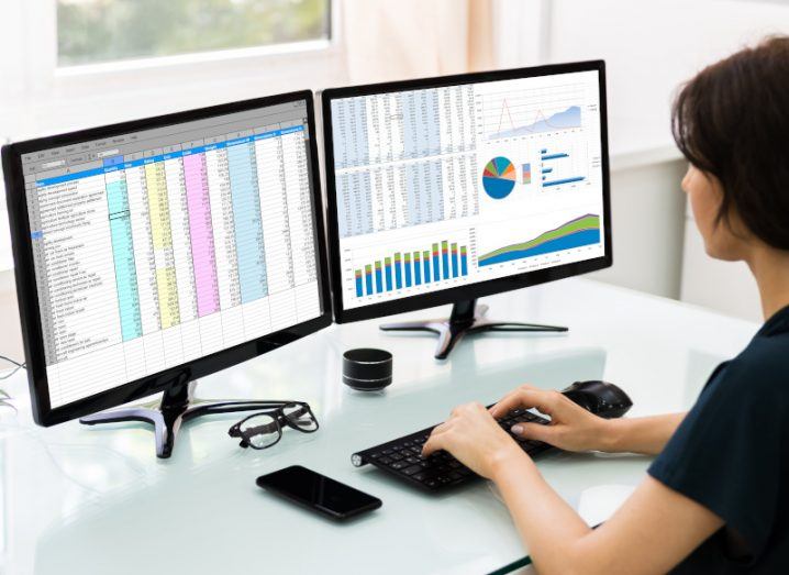 A person typing on a keyboard while looking at two desktop monitors. The monitors both have spreadsheets on them containing various forms of data.