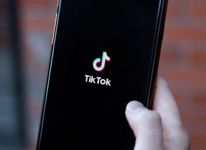 TikTok logo on a mobile phone screen being held in a person's hand. There is a blurry red wall in the background.