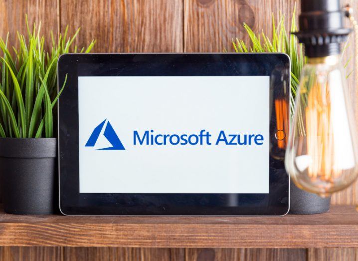 The Microsoft Azure logo on a tablet screen, which is resting on a brown wooden table next to two house plants. A light bulb is visible on the right of the image.