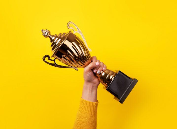 A hand holds a large gold trophy aloft against a sunflower-yellow background.