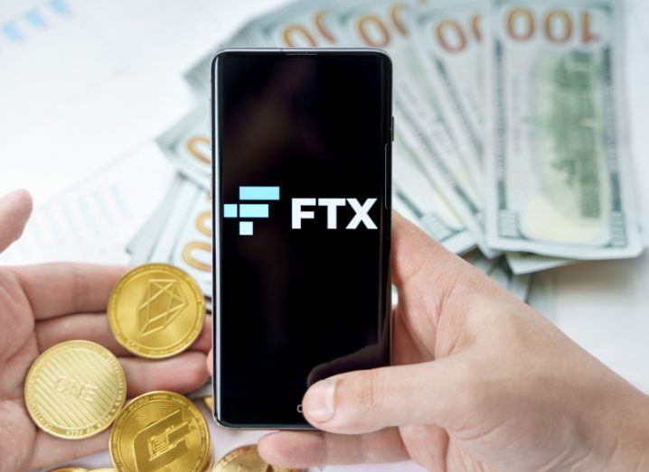 FTX crypto exchange logo on a mobile phone held in a person's hand. There are multiple €100 notes on a table in the background. Another hand is holding a bunch of gold coins that represent cryptocurrencies.