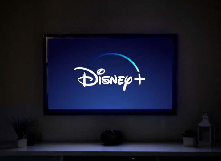 The Disney Plus logo on a TV screen, in a dark room. The light from the TV is reflecting off a table underneath, which has a couple of lamps and small house plants on its surface.