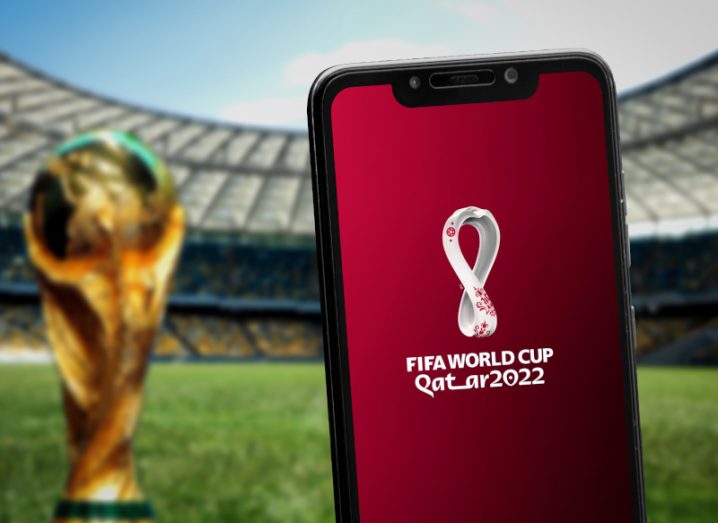 Smartphone with Qatar World Cup advertised on it and a World Cup trophy with a football stadium in the background.