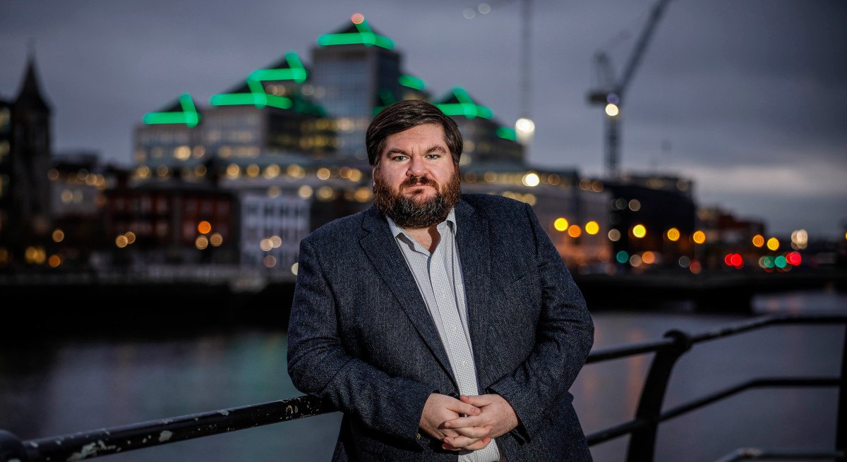 Dr Joe Fitzsimons, founder of Horizon Quantum Computing pictured leaning against a bridge at night with a city skyline behind him and a building with green lights.