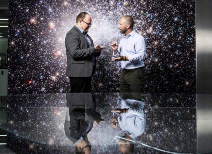 Two men standing together with a space background. The men are reflected on a table next to them. They are the CEOs of Tyndall and Varadis.