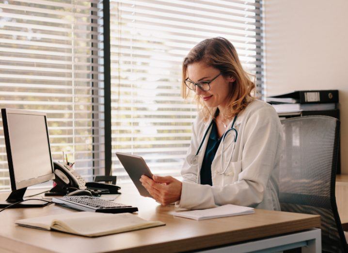A doctor sits at her desk with a tablet in hand. Represents the use of deepfake in medicine and pharma.