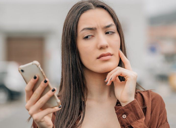 Woman holding a smartphone in hand and looking at it suspiciously. Symbolises spoofing carried out by websites such as iSpoof.