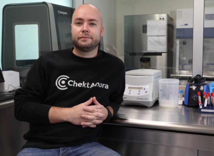 A man wearing a jumper with the ChektAhora logo on it sitting in a lab with testing kits and instruments around him.