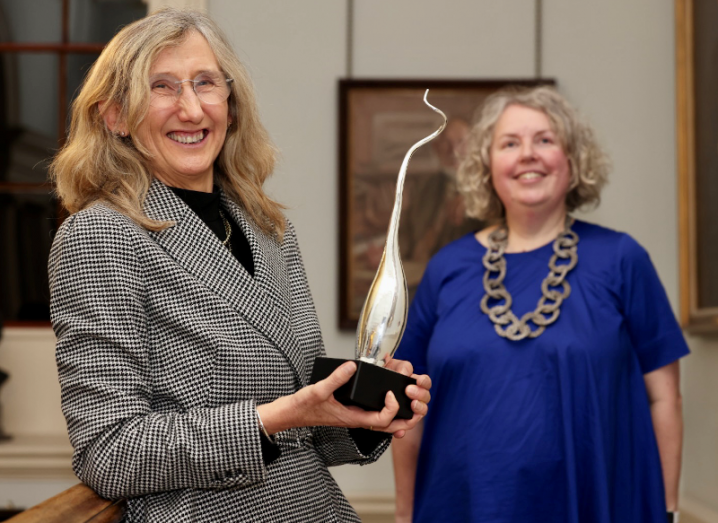 A woman holding a large award statue. Behind her is Dr Linda Doyle, TCD provost.