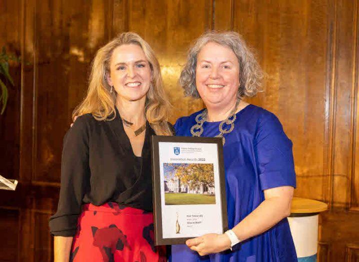 Two women stand on stage smiling at the camera. One is presenting the other with an award.