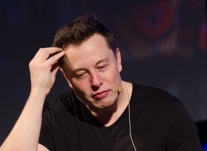Elon Musk sitting at a stage wearing a black t-shirt, with a microphone attached to his face and his right hand on his head.