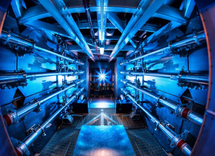 The interior of a nuclear fusion reactor support structure. Pipes are laid across the walls and ceiling of a blue lit hallway, with a bright blue light visible at the end of the hallway.