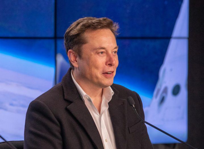 A man sitting in front of a microphone, with screens in the background showing a view of space. He is Elon Musk.