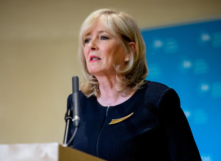 A woman speaking in front of a microphone, with a tan wall with a blue poster in the background. She is EU Ombudsman Dr Emily O'Reilly.